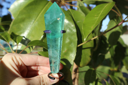 Green fluorite crystal wand held by a hand with leaves in the background